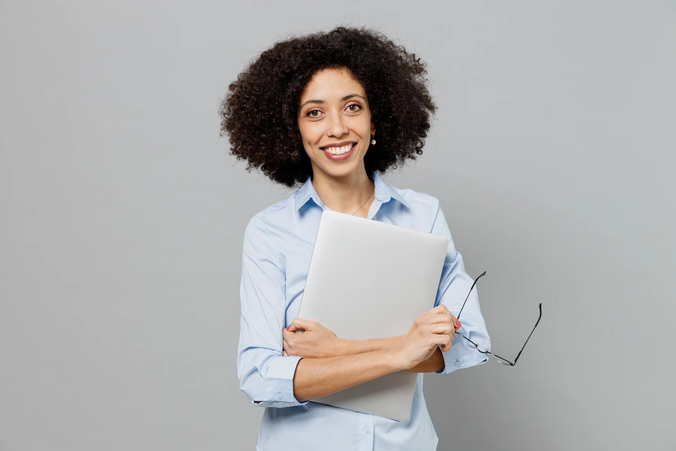 Femme professionnelle souriante en chemise bleue tenant un ordinateur portable fermé et des lunettes, posant dans un environnement de bureau sur fond gris neutre